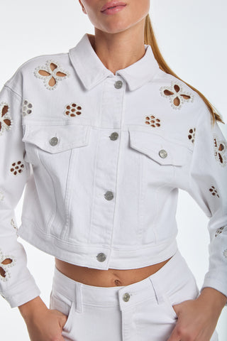 A person wearing a Kaia Pearl Jean Jacket - White and pants. This cropped silhouette, enhanced by embroidered floral cut-outs with beaded flowers on sleeves and shoulders, features silver buttons and is paired with long hair tied back.