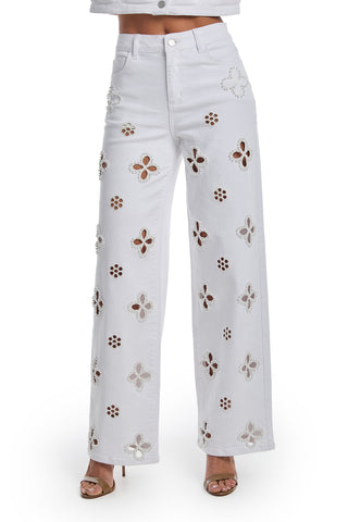 A person wears the Kaia Pearl High Rise Jean - White, featuring straight-leg stretch denim with floral cutouts and pearl embroidery, paired with open-toe heeled sandals and a partially visible cropped white top.