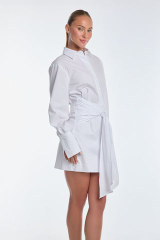 A person with long hair pulled back smiles gently while posing in the Lana Dress - White, a luxurious poplin shirt dress with wrap-tie detailing and an asymmetrical hem, set against a plain white background.