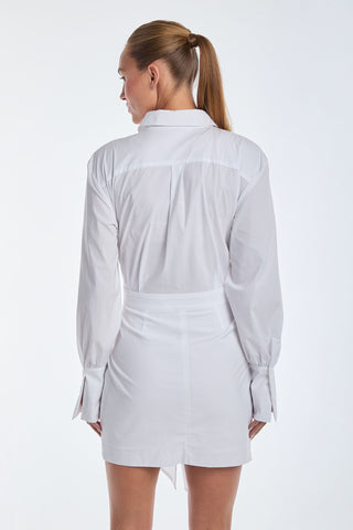 A woman with long hair in a ponytail, wearing the elegant Lana Dress - White, faces away. The luxurious poplin fabric and wrap-tie detailing accentuate the long sleeves and fitted waist against a plain white backdrop.