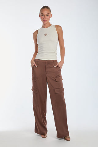 A woman stands against a plain background, wearing the Rosette Tank Top - Pearl and high-waisted brown cargo pants. Her hands in her pockets highlight the rose embroidery detail. She wears sandals, has hair tied back, and offers a slight smile.