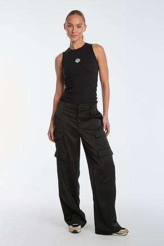 A person confidently poses against a white background wearing the Rosette Tank Top - Black with black cargo pants and stylish black and beige sneakers. They stand casually, slightly bending one leg to showcase their chic outfit.