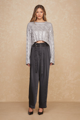 A person wearing a Metallic Cable Knit Sweater in silver, ideal for your fall wardrobe, along with high-waisted black pleated pants featuring a belted waist and black pointed-toe heels, stands against a light textured background.