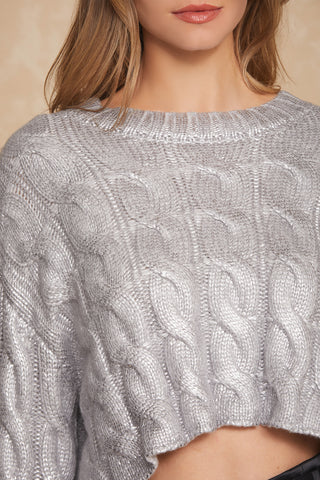 A person with long, wavy hair is wearing a Metallic Cable Knit Sweater in silver, ideal for a Fall wardrobe. The chunky design and cropped cut stylishly reveal part of the midriff against a neutral, soft beige background.