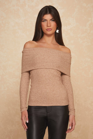 A person with long dark hair wearing an off-the-shoulder Amora Ribbed Knit Top in Praline and sleek black leather pants stands elegantly against a textured beige background. Adorned with statement earrings, they exude sophistication with their neutral expression.