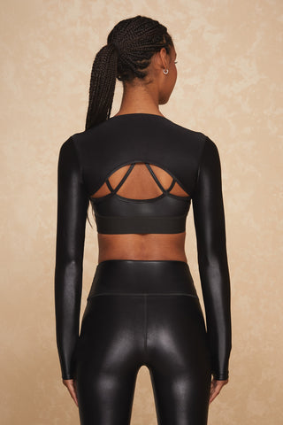 Rear view of a person wearing the Liquid Shrug - Black Gloss, a sleek black long-sleeve bolero top with cut-out details, along with matching leggings. Their hair is styled in a braided ponytail against a subtly textured light beige background.