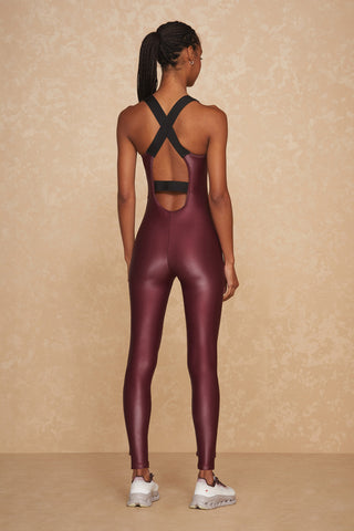 Dressed in the Vixen Jumpsuit - Cherry, designed with a liquid shine fabric and featuring a maroon hue along with a black crisscross back design, an individual confidently stands against a beige textured backdrop, complementing the outfit with white sneakers.