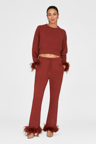 A woman in a rust-colored lounge set with ostrich feather trim on cuffs and hem stands against a white backdrop. Her hair is neatly tied back, and she pairs her Rada Feather Trim Pant - Burgundy with beige heels.