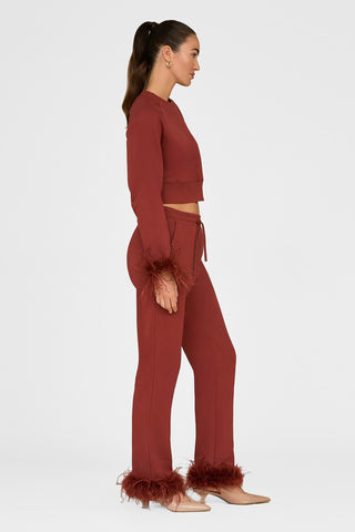 A person is elegantly dressed in a rust-colored outfit with ostrich feather-trimmed sleeves and the Rada Feather Trim Pant in Burgundy. The ensemble includes a cropped top and flared pants, perfectly matched with beige heels. Their long hair is styled in a ponytail, completing the chic look.