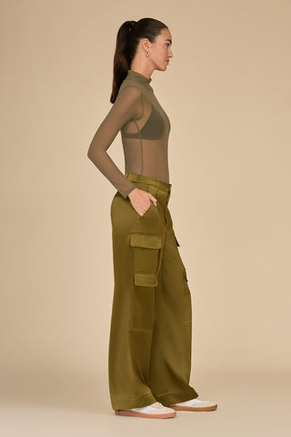 A person stands in profile against a neutral background, wearing a sheer olive top paired with the Milan Satin Cargo Pant - Olive, which features mid-rise cargo details and roomy pockets. Their hair is styled in a ponytail, and they complete the outfit with shoes that have a white base and light sole.