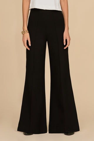 A person dressed in the high-waisted, black Ava Ponte Wide Leg Pant poses against a beige background. They complement it seamlessly with a white top and embellish their left wrist with a gold bracelet, while their hands hang naturally by their sides.