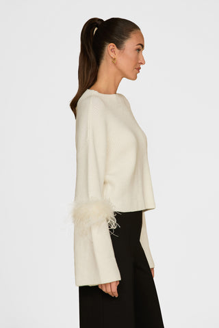 A person with long brown hair in a ponytail wears the Bella Feather Trim Sweater - Pearl, featuring luxurious knit and feather accents on the sleeves, paired with black pants against a plain white background.