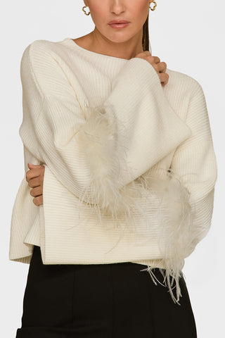 A woman in a Bella Feather Trim Sweater - Pearl with feather trim sleeves and black pants holds one arm across her body. Her gold hoop earrings glint against the light gray backdrop, complementing the chic sophistication of her luxurious knit sweater.