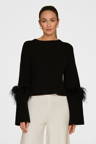 A person stands in the elegant Bella Feather Trim Sweater - Black, showcasing detailed feather accents on the sleeves, paired with cream pants. Their long hair is tied back and gold hoop earrings complete the look against a simple white backdrop.