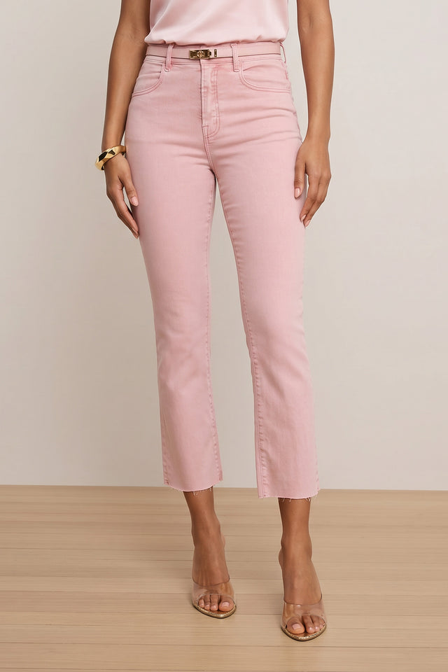 A woman wears the Nicolette Cropped Jean - Blush with a matching light pink top, clear-strap heeled sandals, and a gold bracelet, standing on a light wood floor against a beige background. Made from Four-Way Stretch Denim for comfort.