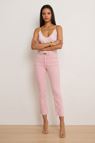 A woman stands with arms crossed, wearing a light pink camisole, Nicolette Cropped Jean - Blush made from comfortable Four-Way Stretch Denim, clear heeled sandals, and gold jewelry against a plain light background.