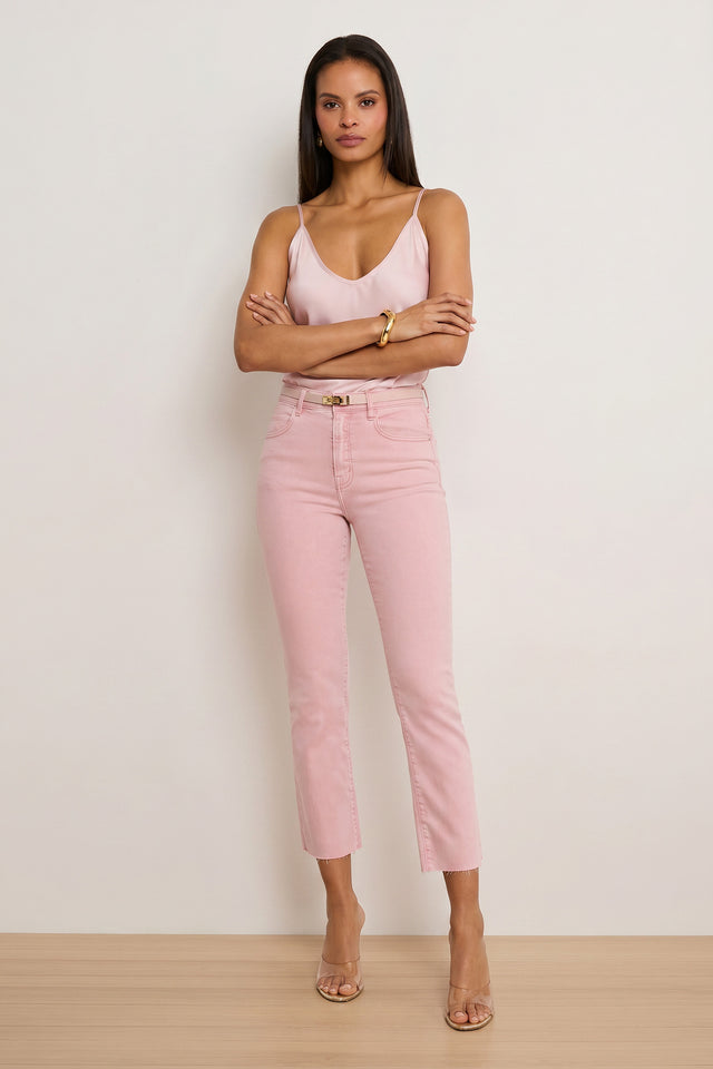A woman stands with arms crossed, wearing a light pink camisole, Nicolette Cropped Jean - Blush made from comfortable Four-Way Stretch Denim, clear heeled sandals, and gold jewelry against a plain light background.
