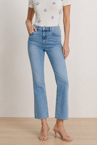 A woman stands against a plain background on a wooden floor, wearing the Nicolette Cropped Jean in medium blue with a straight-leg fit, paired with a white short-sleeve shirt featuring blue anchor prints and beige heeled sandals.
