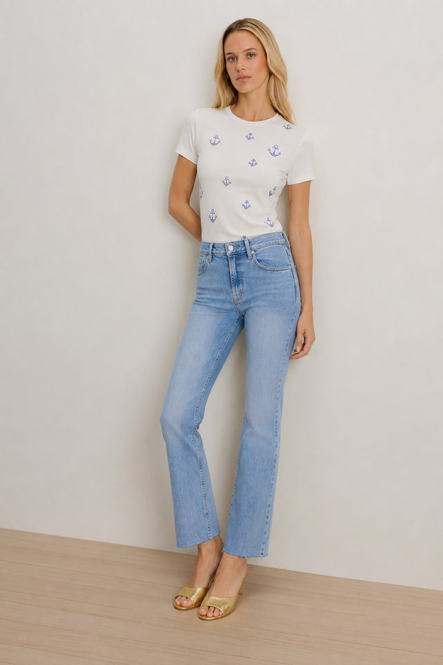 A woman with long blonde hair stands against a plain wall in a white t-shirt with blue anchor prints, gold open-toe heels, and the Nicolette Cropped Jean - Medium Blue, featuring mid-to-high rise and four-way stretch denim.