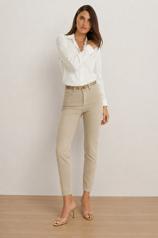 A woman stands on a wooden floor wearing a white blouse, the Nina Cropped Skinny Jean in oatmeal, and clear heeled sandals. She has long brown hair and is gazing at the camera with one hand touching her collar.