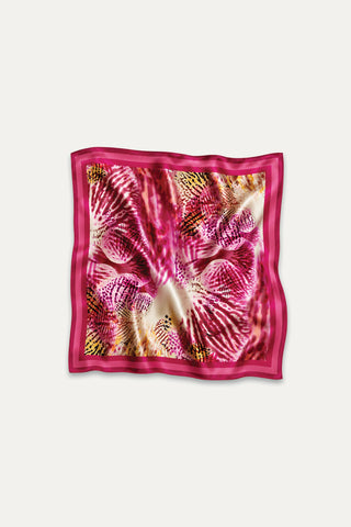 The Printed Satin Scarf - Bright Orchid features a square, silky design with vibrant pink, red, and yellow abstract feather-like patterns reminiscent of orchids, accented by a solid pink border and shown laid flat with gentle folds.