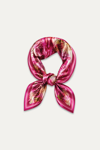 The Printed Satin Scarf - Bright Orchid features a vibrant pink, red, and gold abstract pattern. Silky and versatile, it’s styled in a loose knot on a white background—perfect for adding bold flair as a headscarf.