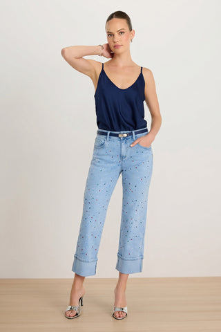 A woman stands on a wood floor wearing the Ren Cropped Cuff Jean - Confetti with small colorful embellishments, a navy camisole, silver heeled sandals, and a belt. One hand is in her pocket and the other touches her head.