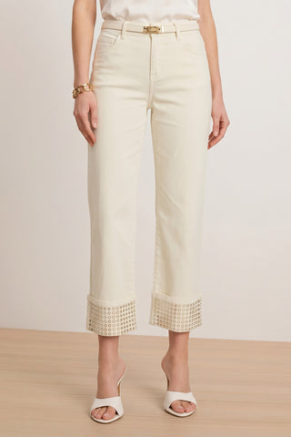 A woman stands on a wooden floor wearing the Ren Cropped Cuff Jean - Gold Mirror with cut-out cuff details, paired with a white top, white heeled sandals, and a gold bracelet.