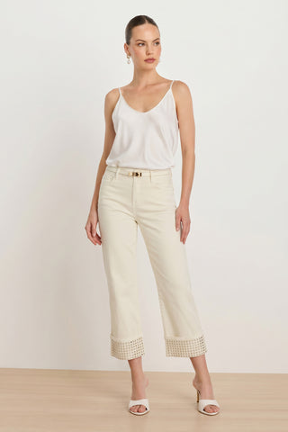 A woman models the Ren Cropped Cuff Jean - Gold Mirror, paired with a white sleeveless top, white heeled sandals, and hoop earrings against a plain light background.