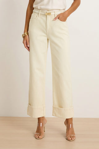 A woman wears the Ren Cropped Cuff Jean in Ivory with a white top, clear-strap heeled sandals, gold bangles, and a thin belt, standing on a light wood floor against a plain background.