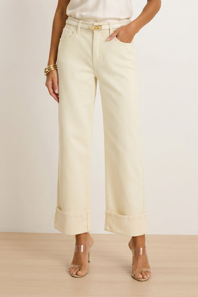 A woman wears the Ren Cropped Cuff Jean in Ivory with a white top, clear-strap heeled sandals, gold bangles, and a thin belt, standing on a light wood floor against a plain background.