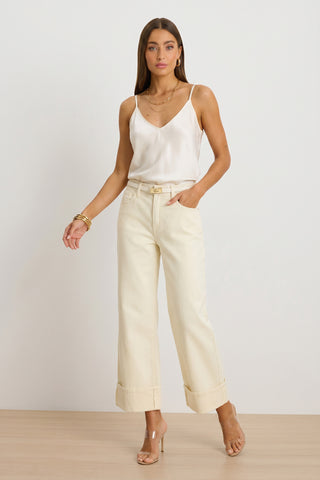 A woman stands against a plain background wearing a white satin camisole, the Ren Cropped Cuff Jean in Ivory with cuffed hems and four-way stretch, nude high-heeled sandals, layered necklaces, and gold bangles.