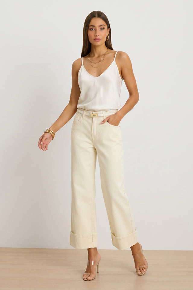 A woman stands against a plain background wearing a white satin camisole, the Ren Cropped Cuff Jean in Ivory with cuffed hems and four-way stretch, nude high-heeled sandals, layered necklaces, and gold bangles.