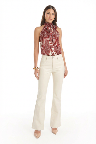 A woman stands against a white background wearing the Rina Tie Neck Blouse in Garnet Crush, paired with cream flare pants, nude heels, and gold bracelets.