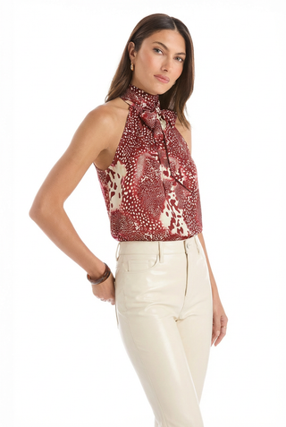 A woman with long, straight brown hair and a neutral expression wears the Rina Tie Neck Blouse - Garnet Crush, a sleeveless red and white top with a bow at the neck, paired with high-waisted cream pants against a white background.