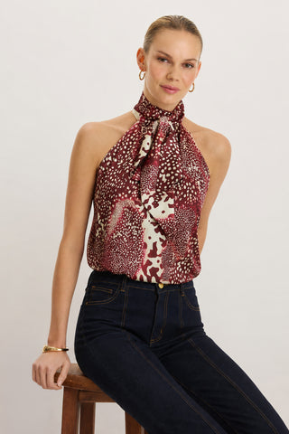 A woman wearing the Rina Tie Neck Blouse in Garnet Crush, styled with dark jeans, gold hoop earrings, and a bracelet, sits on a wooden stool against a plain white background.