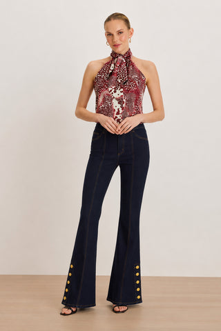 A woman stands on a wooden floor wearing the Rina Tie Neck Blouse in Garnet Crush, styled as a red patterned halter top, paired with high-waisted dark flared jeans featuring gold buttons at the ankles, black heeled sandals, and small earrings, set against a plain light background.