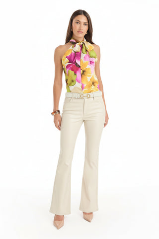 A woman with long brown hair and a neutral expression faces forward, wearing the Rina Tie Neck Blouse in floral print with large yellow, green, and pink flowers, cream-colored flared pants, and pointed nude heels.