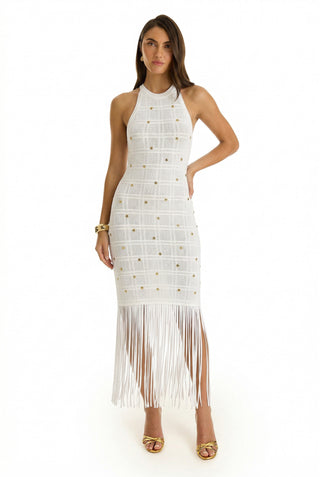 A woman stands in the Romy Embellished Dress - Pearl, with a halter neckline, gold accents, fringe hem, and metallic open-toe heels—an elegant pick for summer events. She poses against a plain white background.