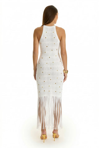 A woman faces away in the Romy Embellished Dress - Pearl, a sleeveless white halter style with gold accents, grid pattern, and long fringe hem. She pairs it with yellow heels and a bracelet—ideal for summer soirées.
