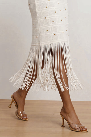 A person walks on a wooden floor wearing the Romy Embellished Dress - Pearl, featuring fringe at the hem and gold studs, paired with transparent high-heeled sandals with gold soles for an elegant look.