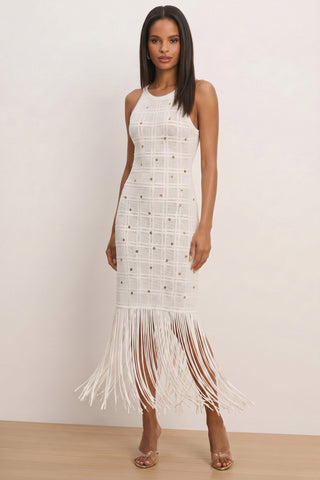 A woman stands indoors wearing the Romy Embellished Dress - Pearl, featuring a grid pattern with gold accents, a halter neckline, and a long fringed hem. She pairs it with clear high-heeled sandals and has straight hair.