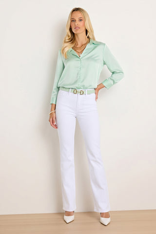 A woman stands against a plain light-colored wall, wearing the Sara Silk Blouse - Mint, white high-waisted pants with a matching belt, white heels, and layered gold necklaces.