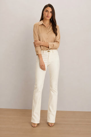 A woman stands against a plain background wearing the Sara Silk Blouse in Mocha—a timeless women's wardrobe staple—paired with white high-waisted pants and beige heels. She has long brown hair, a neutral expression, and one arm bent resting near her waist, perfectly highlighting the elegant silk blouse.