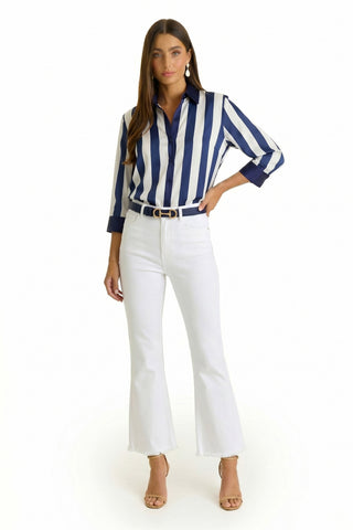 A woman faces forward in the Sara Silk Blouse - Navy Stripe, featuring blue and white vertical stripes with three-quarter sleeves, paired with white flared jeans, a black belt, and beige high-heeled sandals against a plain white background.