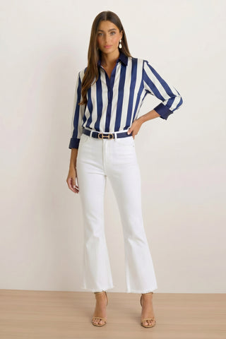 A woman stands against a plain background, wearing the Sara Silk Blouse - Navy Stripe, white high-waisted flared jeans, a black belt, beige heeled sandals, gold earrings, and her long hair flowing.