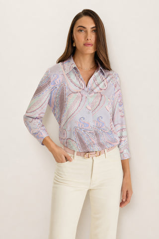 A woman models the Sara Silk Blouse – Pastel Paisley, featuring long sleeves and a silky finish, paired with cream-colored pants. She stands with one hand in her pocket against a plain light background.