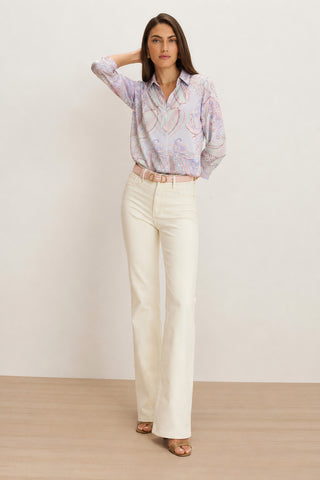 A woman stands on a wood floor wearing the Sara Silk Blouse in Pastel Paisley, tucked into high-waisted white pants with a belt and heeled sandals.