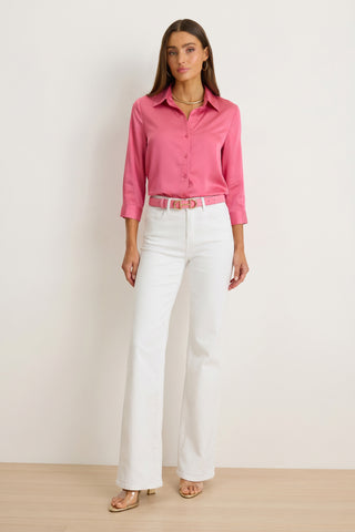 A woman stands by a plain light wall, wearing the Sara Silk Blouse in petal pink with three-quarter sleeves, white high-waisted wide-leg pants, a matching pink belt, and beige heeled sandals.