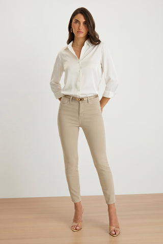 A woman with long dark hair and a neutral expression poses indoors against a plain background, wearing the Sara Silk Blouse - White paired with beige fitted pants, a belt, and open-toe heels.
